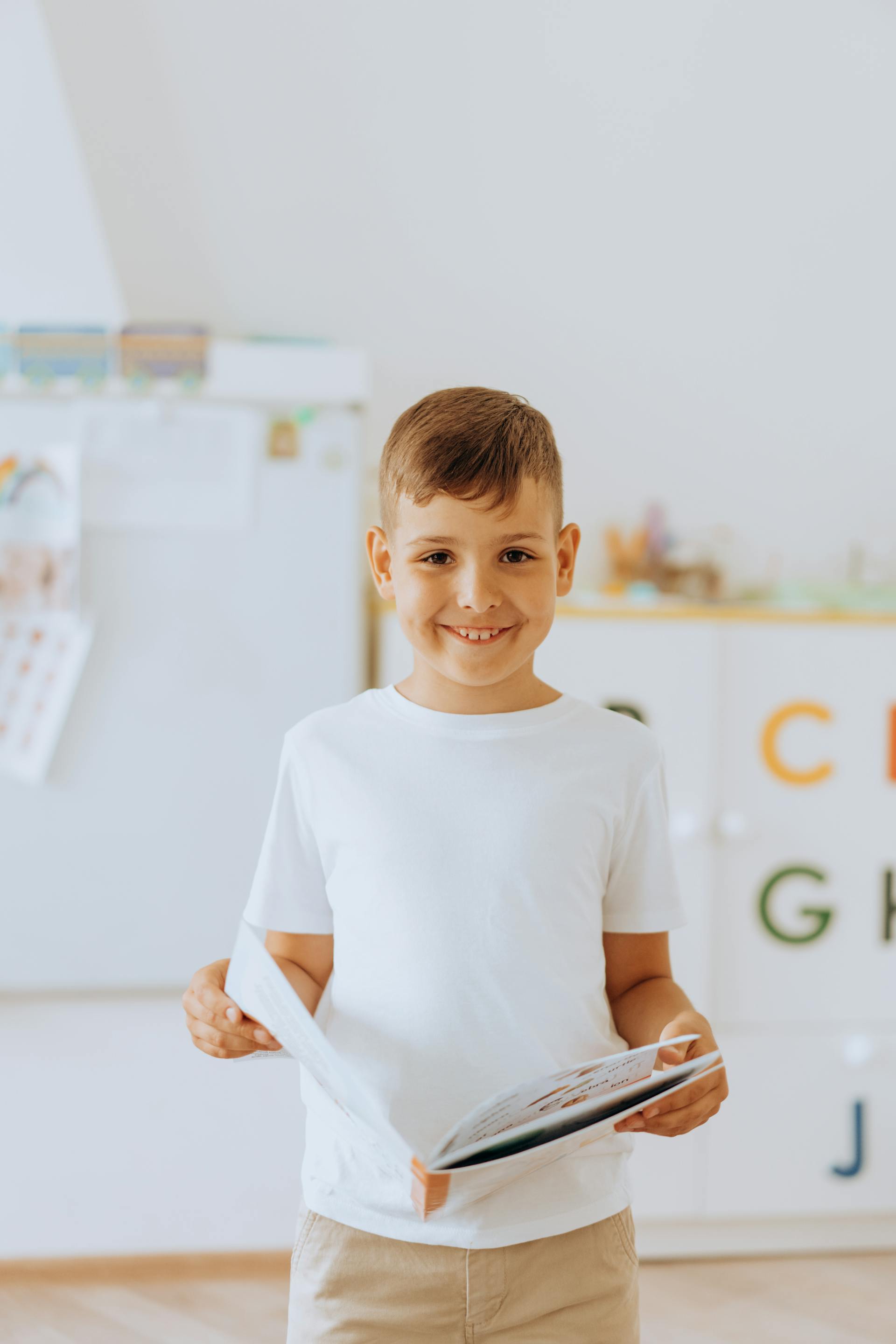 A smiling boy holding a book in a bright classroom